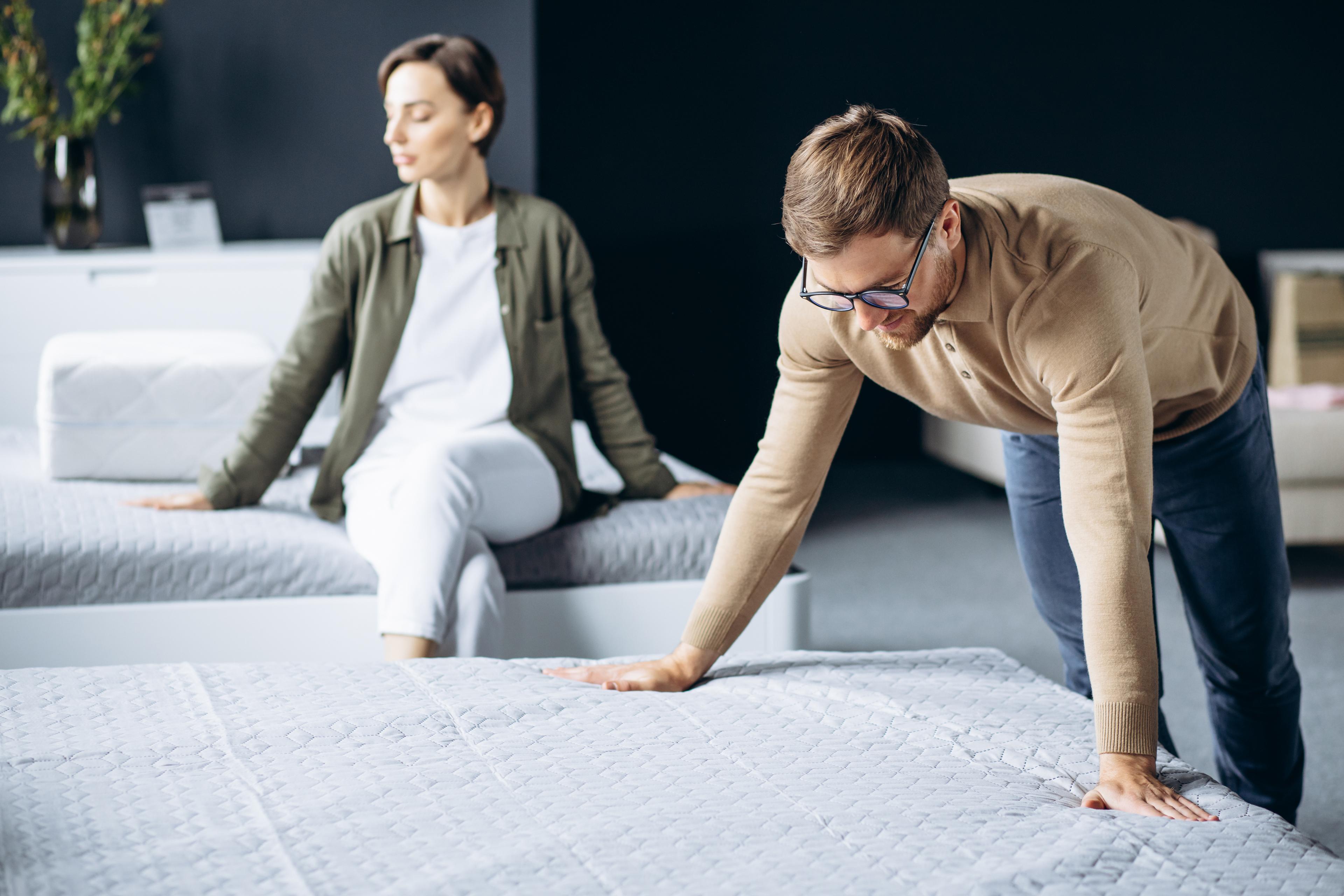 A man and woman test mattresses in a bright store. He leans forward, both hands on the mattress to test the firmness while the woman is sitting on another mattress to test the comfort. They are not engaged, perhaps zoned in their own mattress experiences.